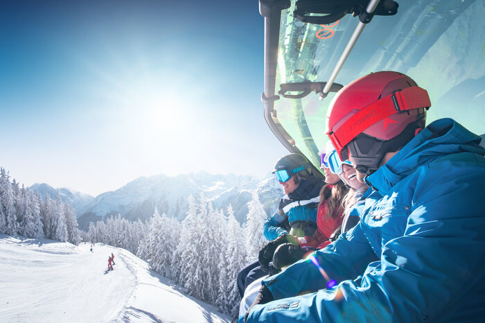 Skiers on Chairlift with Panoramic View | © Ski amadé Group of skiers wearing helmets and goggles rides a chairlift with weather cover over a snow-covered mountain landscape. | © Ski amadé