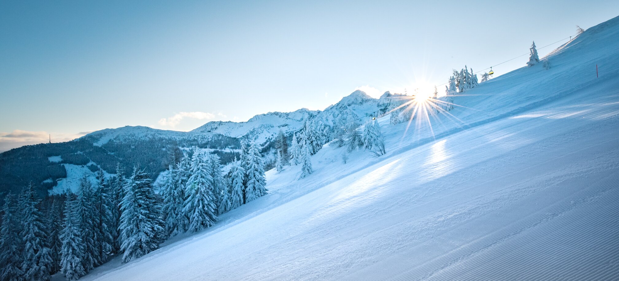 Shining sun over a groomed ski slope with snow-covered trees. | © Ski amadé