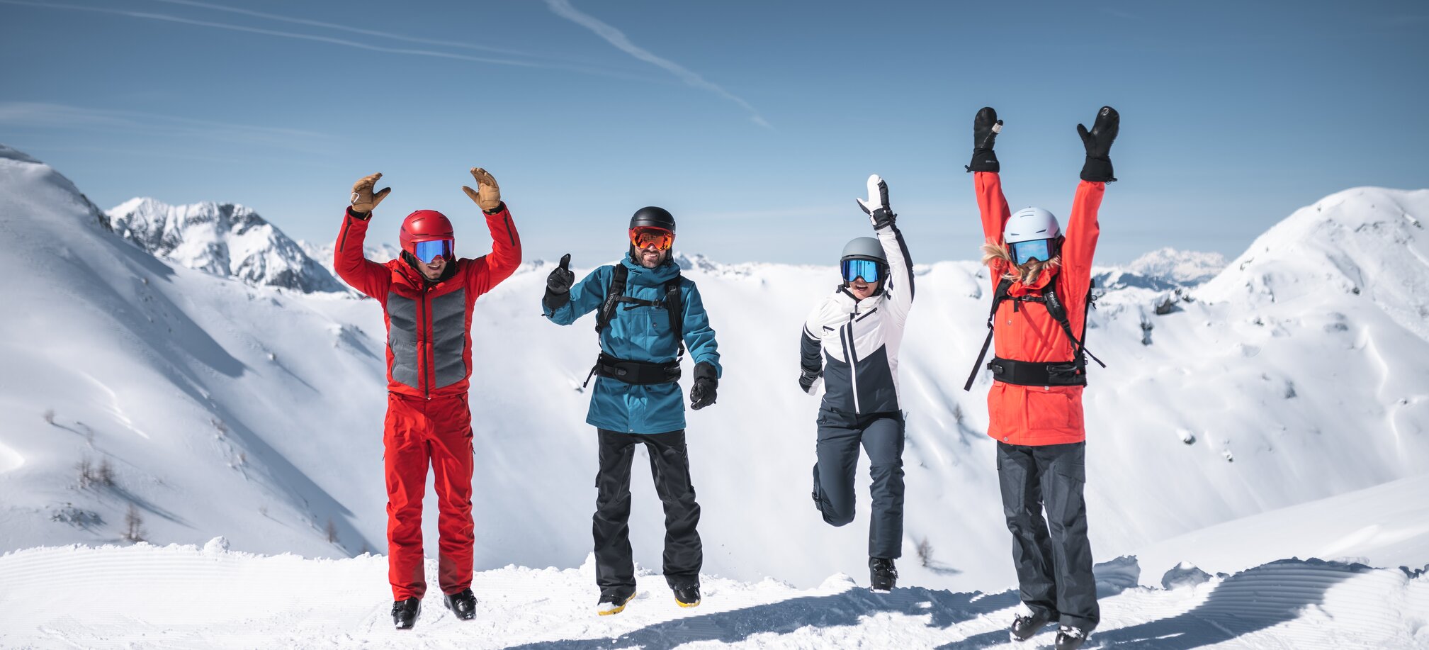 Four skiers jump in the air together with snowy mountain peaks and sunlight in the background