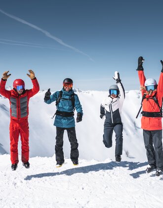 Four skiers jump in the air together with snowy mountain peaks and sunlight in the background