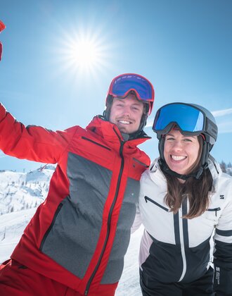 Ein Skifahrer und eine Skifahrerin posieren lachend bei Sonnenschein auf der Skipiste