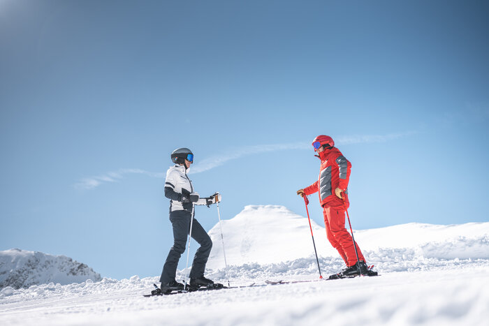 Zwei Skifahrer im Gespräch auf sonniger Piste im Gebirge Zwei Skifahrer stehen sich auf schneebedeckter Piste gegenüber, umgeben von Bergen und blauem Himmel.