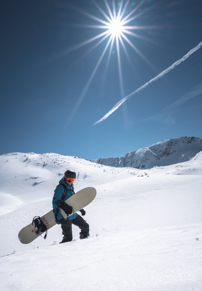 Snowboarder steigt bei Sonnenschein durch Tiefschnee Snowboarder mit Brett in der Hand steigt durch unberührten Tiefschnee, über ihm strahlt die Sonne am Himmel.