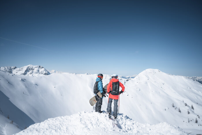 Skier and snowboarder enjoy summit panorama Two winter sports enthusiasts stand on a snowy summit, overlooking a vast panorama of white mountains and ridges.