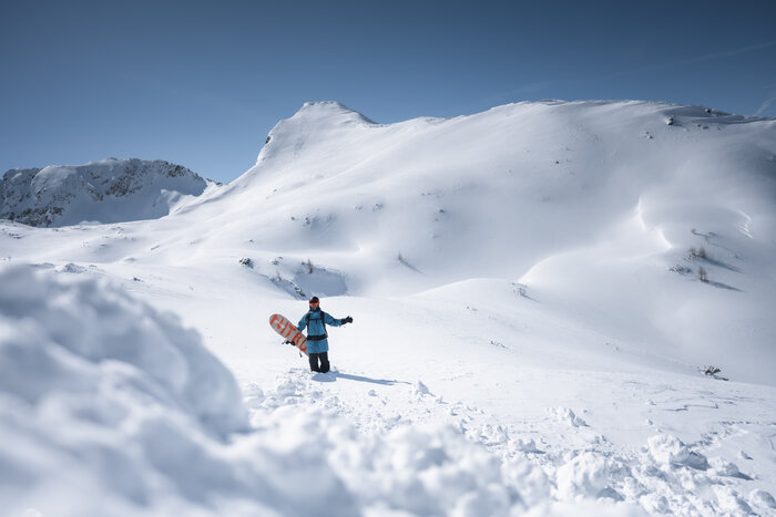Snowboarder walking in untouched winter landscape A snowboarder with board in deep snow, wide white slopes ahead and a snow-covered mountain in the background.