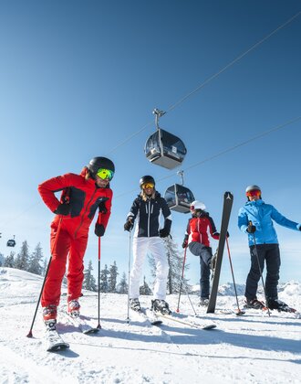 Skifahrer in bunten Skianzügen stehen auf der Piste und über ihnen schwebt die Gondel und eine hat einen Ski angehoben. | © Ski amadé
