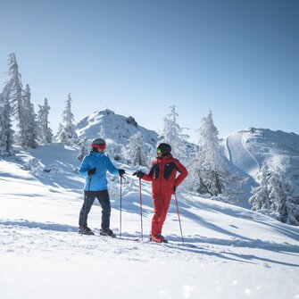 Zwei Männer in rotem und blauem Skianzug unterhalten sich auf der Piste, mit schöner Schneelanfschaft im Hintergrund | © Ski amadé