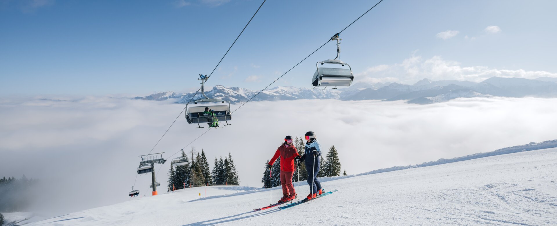 Zwei Skifahrer auf sonniger Piste unter Sessellift mit Blick auf Wolkenmeer und Berge in Ski amadé | © Ski amadé