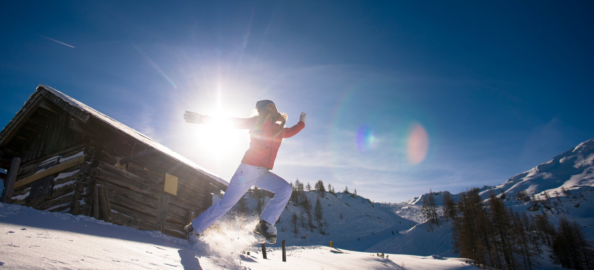 A female skier jumps through the snow in front of a wooden hut in the sun. | © Ski amadé
