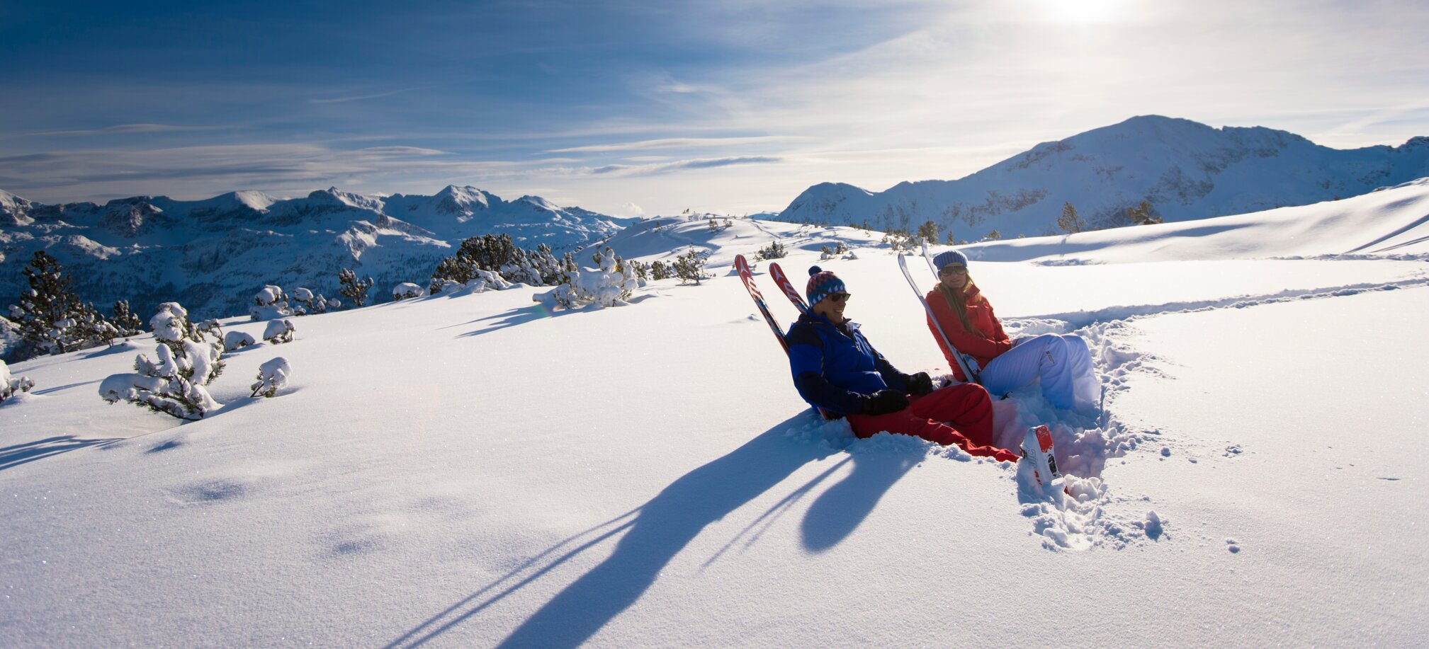 Zwei Skifahrer sitzen im tiefen Schnee mit Skiern und genießen die Sonne.