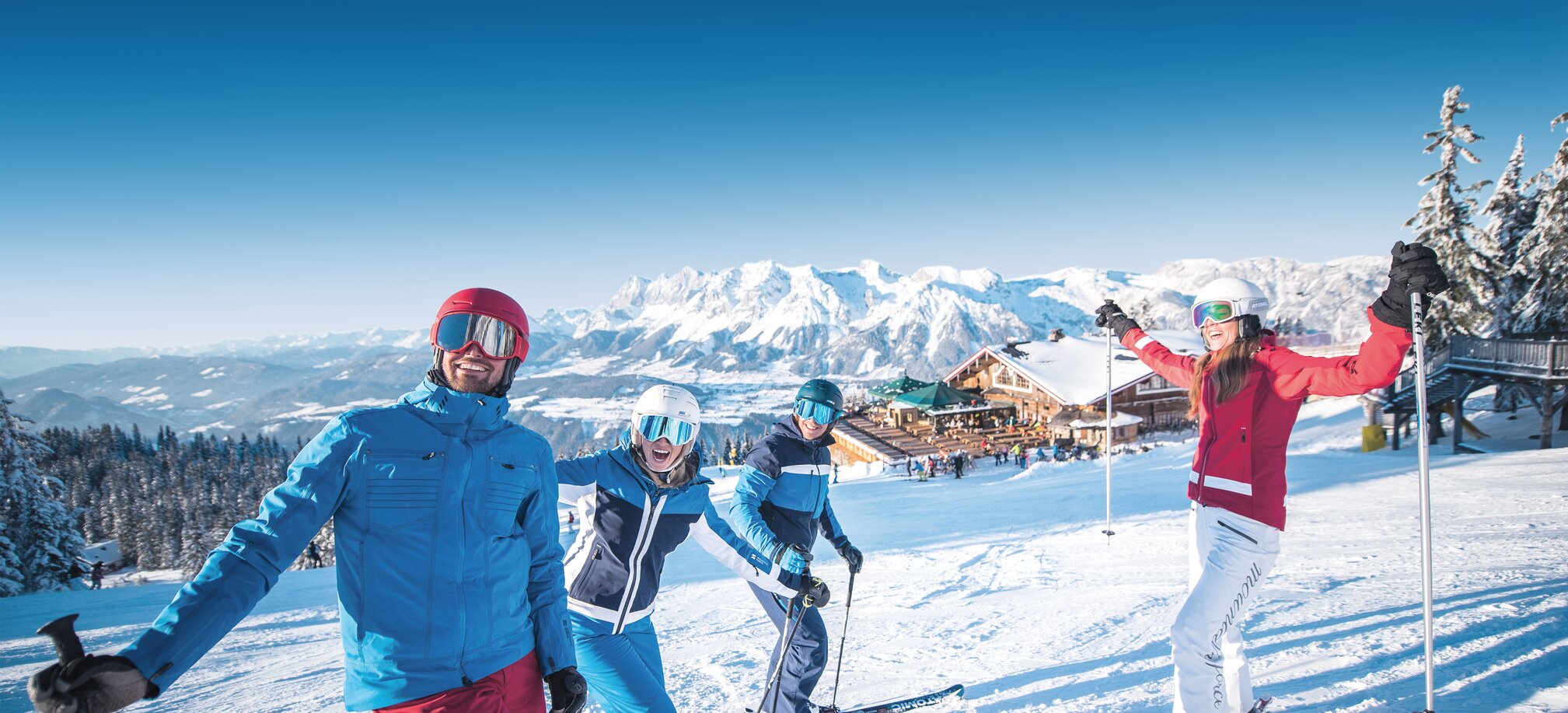 Vier Skifahrer lachen auf der Piste mit einer Berghütte im Hintergrund. | © Ski amadé