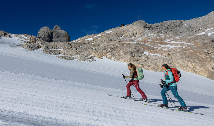 Große Mitterstein-Tour im Anstieg auf sonniger Schneefläche | © Harald Steiner Zwei Frauen steigen mit Tourenski einen verschneiten Hang unter Felswänden hinauf | © Harald Steiner