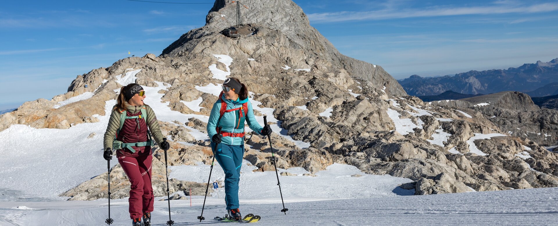 Zwei Frauen gehen mit Skitouren-Ausrüstung vor felsigem Gipfel bei Sonnenschein | © H. Steiner