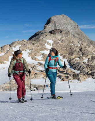 Zwei Frauen gehen mit Skitouren-Ausrüstung vor felsigem Gipfel bei Sonnenschein | © H. Steiner