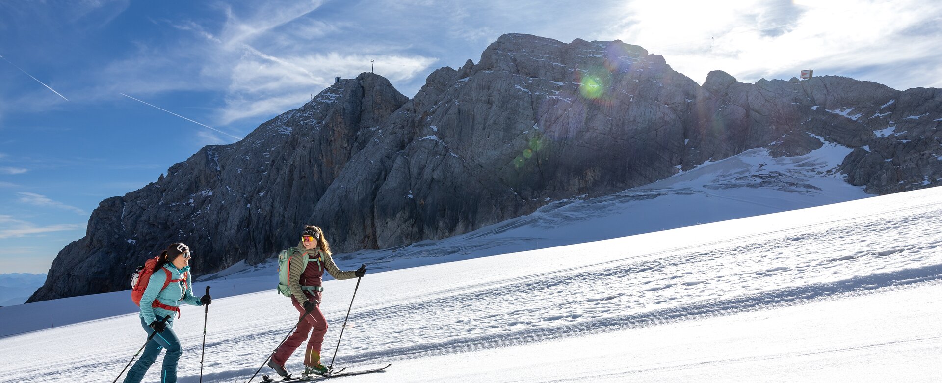 Zwei Frauen steigen mit Tourenski bei Sonne vor steiler Felswand im Schnee bergauf | © Harald Steiner