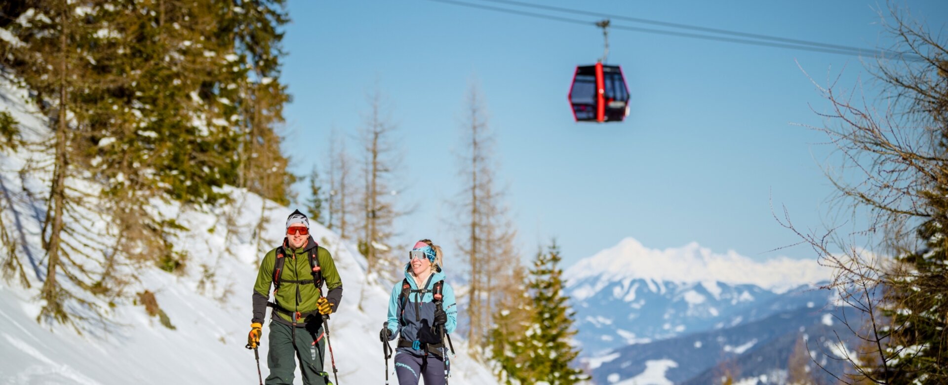 Zwei Skitourengeher steigen eine verschneite Piste am Galsterberg hinauf, während eine rote Gondel über ihnen schwebt. | © Christoph Huber
