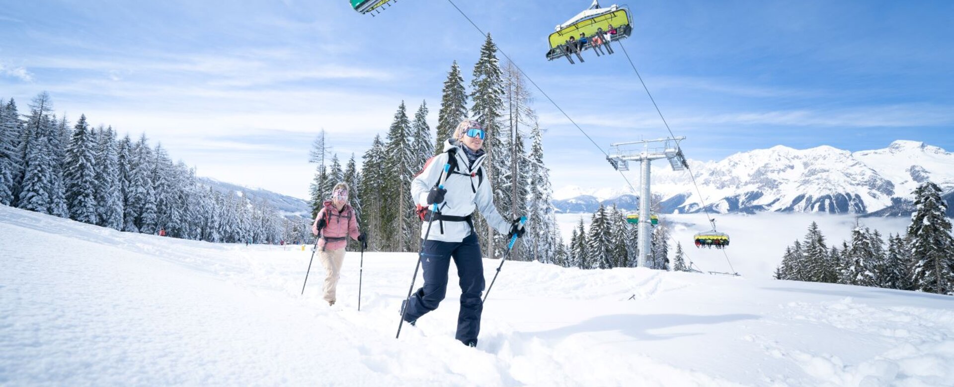 Zwei Skitourengeherinnen steigen durch den Schnee, während über ihnen ein Sessellift mit Skifahrern fährt. | © Josh Absenger