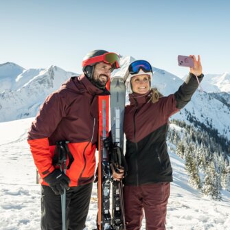 Paar in Skikleidung macht Selfie mit Smartphone vor verschneiter Bergkulisse im Skigebiet Großarltal | © TVB Großarltal - Lorenz Masser