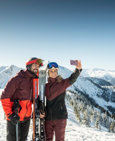 Paar in Skikleidung macht Selfie mit Smartphone vor verschneiter Bergkulisse im Skigebiet Großarltal | © TVB Großarltal - Lorenz Masser