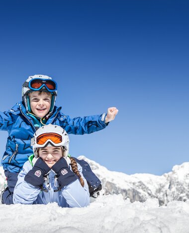 Two children in ski gear playing in the snow with snowy Alpine mountains behind | © Felsch Fotodesign