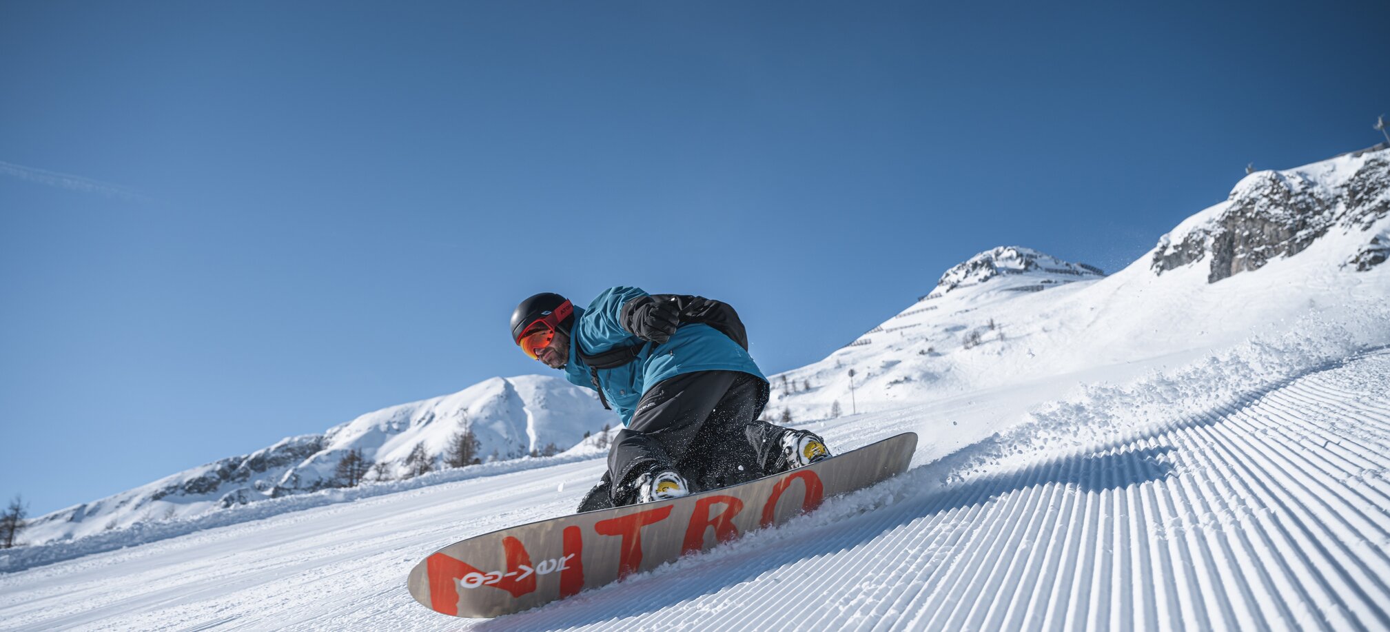 Ein Snowboarder legt sich dynamisch in die Kurve auf präparierter Piste, im Hintergrund verschneite Bergwelt.