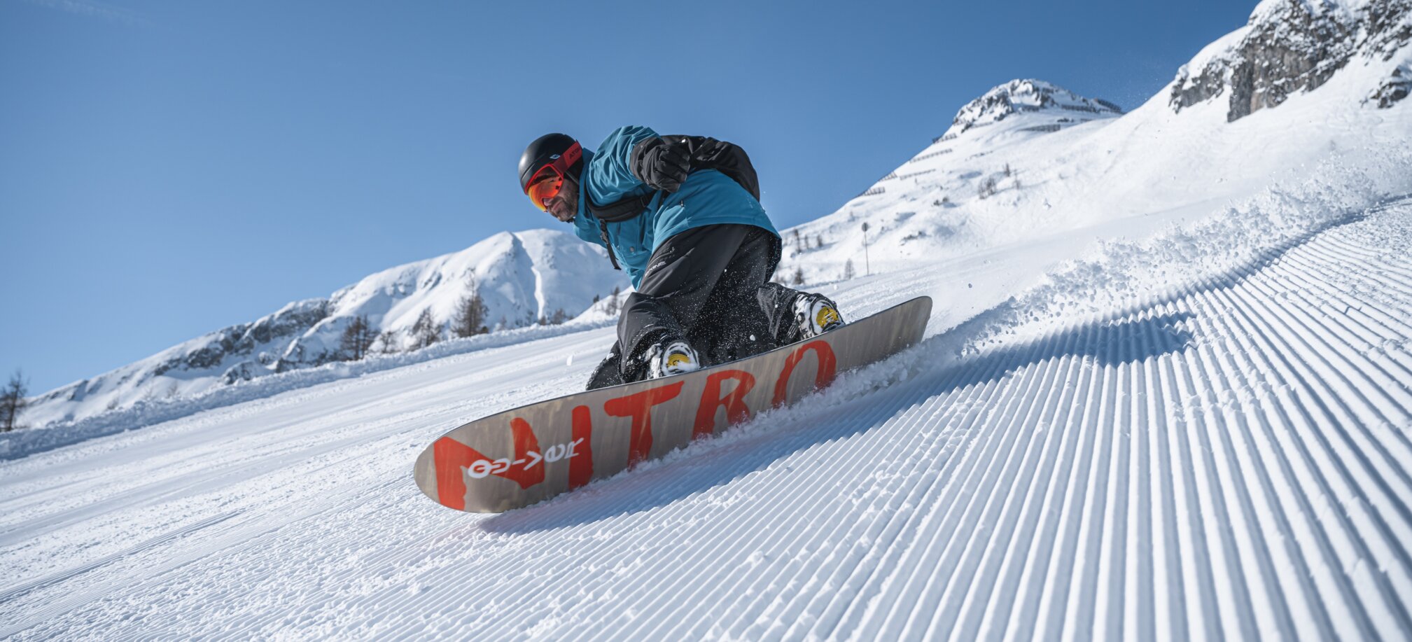 Ein Snowboarder legt sich dynamisch in die Kurve auf präparierter Piste, im Hintergrund verschneite Bergwelt.