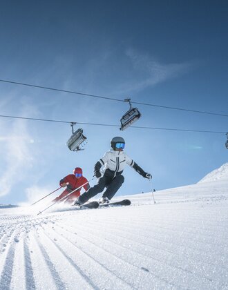 Two skiers ride side by side on groomed slope, above them a chairlift and bright sun shining through a clear sky.