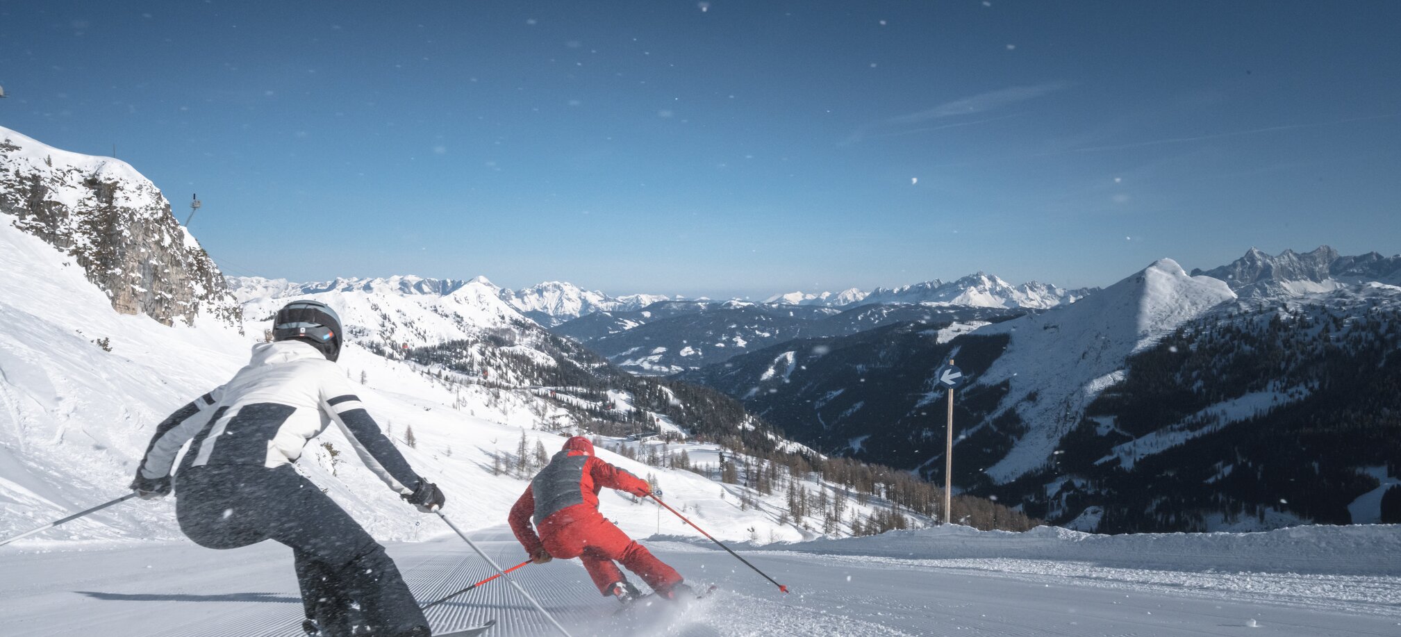 Zwei Skifahrer fahren dynamisch bergab auf perfekt präparierter Piste, Schneestaub fliegt durch die Luft.