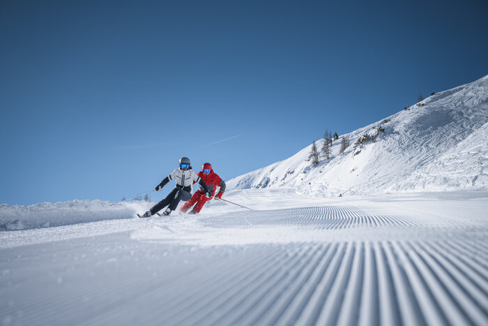 Two skiers carving in sync on perfectly groomed slope Two skiers carve side by side on a freshly groomed slope under a deep blue sky with perfect winter conditions.