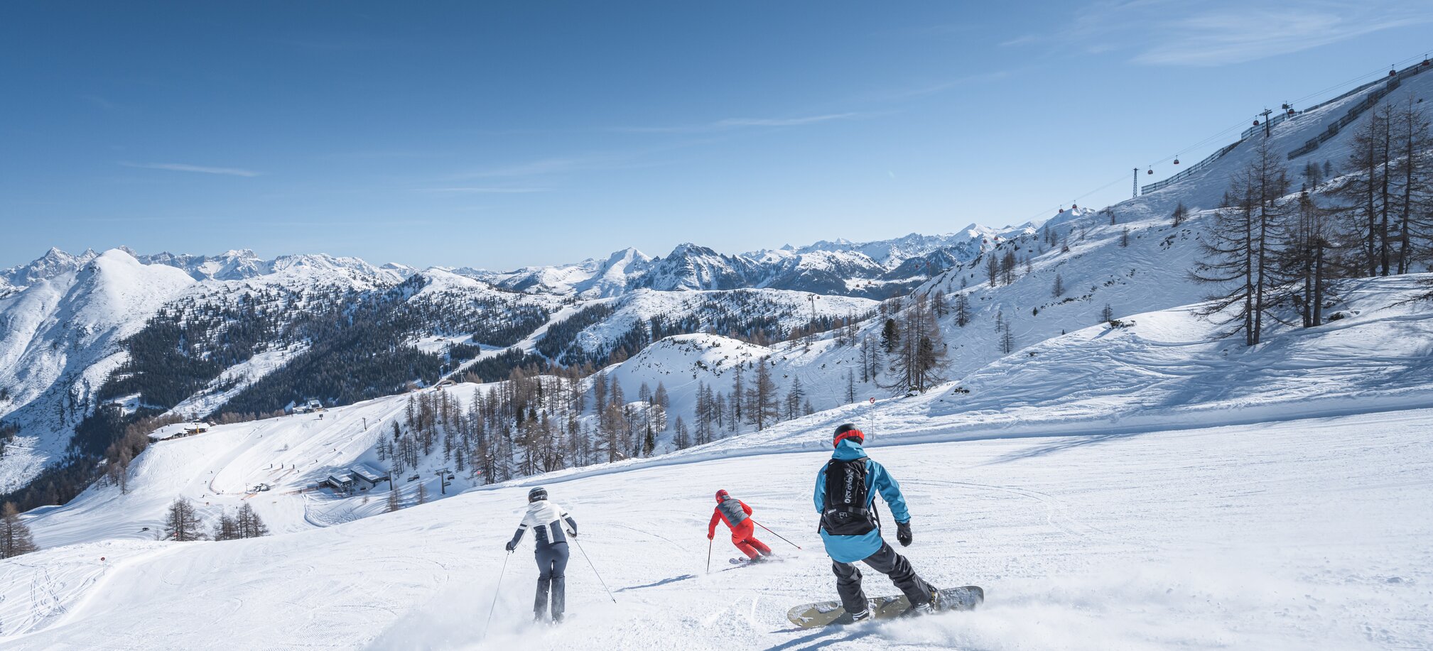 Two skiers and a snowboarder ride together on a wide slope with snowy mountain scenery in the background.