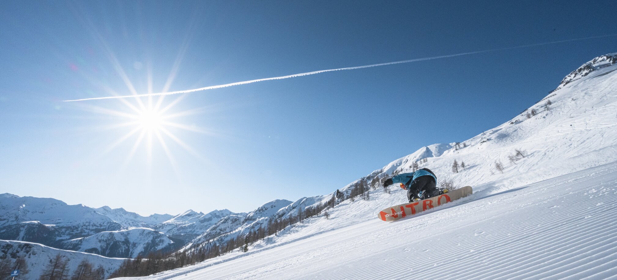 A snowboarder carves a turn on a freshly groomed slope, with the sun shining above and mountains in the background.