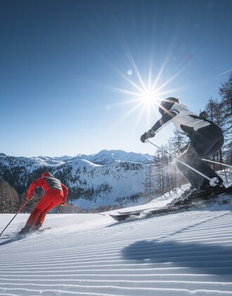 Two skiers carve down a freshly groomed slope, sunbeams shining through trees and above the mountain panorama.