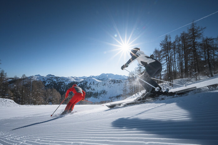 Zwei Skifahrer im Carving-Schwung bei tiefstehender Sonne Zwei Skifahrer carven über frisch präparierte Piste, Sonnenstrahlen leuchten durch Bäume und über Berge.
