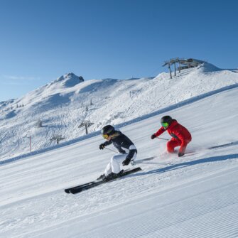 Two skiers carving gracefully on freshly groomed slopes with a stunning mountain backdrop in Ski amadé. Ideal conditions for unforgettable ski days.