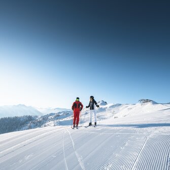 Zwei Skifahrer stehen nebeneinander auf frisch präparierter Skipiste mit Sonne, Sessellift und Alpenblick