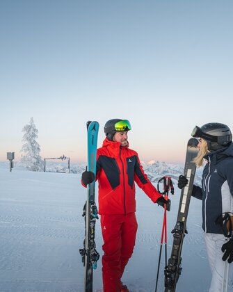 Zwei Skifahrer stehen auf einer verschneiten Piste bei Sonnenaufgang, mit Blick auf eine winterliche Berglandschaft, der Mann trägt einen roten Skianzug, die Frau einen blauen.