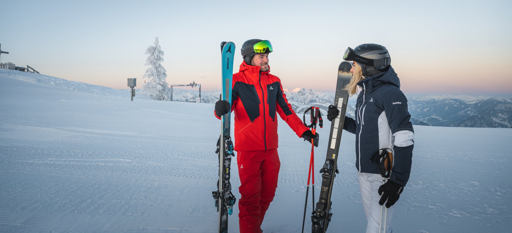 Zwei Skifahrer stehen auf einer verschneiten Piste bei Sonnenaufgang, mit Blick auf eine winterliche Berglandschaft, der Mann trägt einen roten Skianzug, die Frau einen blauen.