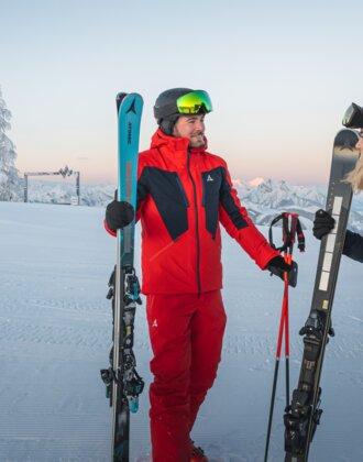 Zwei Skifahrer stehen auf einer verschneiten Piste bei Sonnenaufgang, mit Blick auf eine winterliche Berglandschaft, der Mann trägt einen roten Skianzug, die Frau einen blauen.