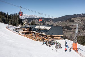 Skihütte mit Sonnenterrasse an einer Skipiste, darüber rote Gondeln und dahinter schneebedeckte Berge