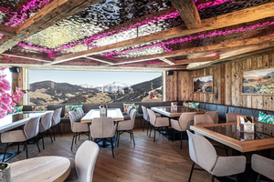 Rustikaler Gastraum mit Holzwänden, Tischen, Sesseln, pink beleuchteter Decke und Bergblick durchs Panoramafenster