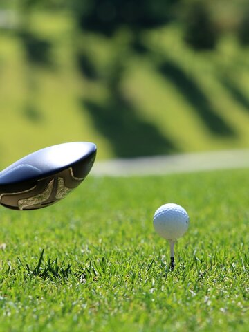 Ein Golfball steht auf einem Tee, kurz vor dem Abschlag mit einem Driver, im Hintergrund ein sonniges Green.
