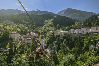 Woman glides on the Flying Waters zipline over rooftops of Bad Gastein, surrounded by forested hills and alpine peaks | © Max Steinbauer Photography