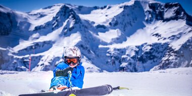 Kind in blauer Skibekleidung sitzt lachend im Schnee vor steilen Bergen in Gastein | © Max Steinbauer