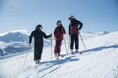Drei Personen in Skiausrüstung stehen auf einer Piste und unterhalten sich lächelnd | © Gasteinertal Tourismus GmbH, Christoph Oberschneider