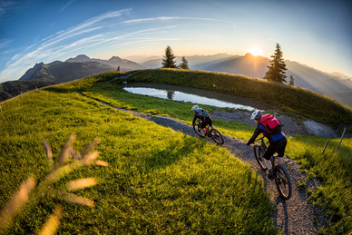 Two mountain bikers ride a narrow trail through green meadows at sunset with scenic views of the Grossarl Valley. | © mtb_travel