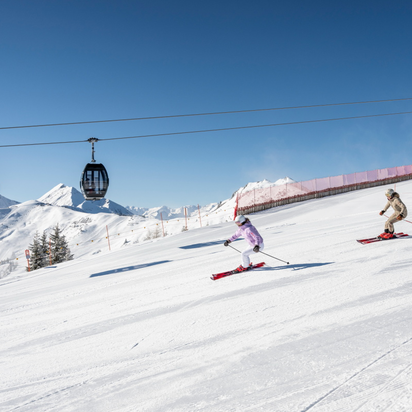 Zwei Skifahrer fahren auf präparierter Piste im Großarltal unter Gondelbahn mit verschneiten Bergen und blauem Himmel | © Lorenz Masser