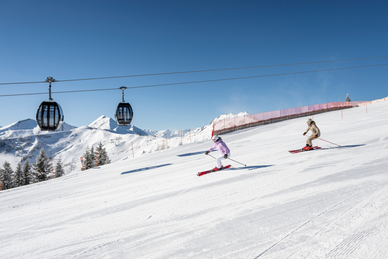 Zwei Skifahrer fahren auf präparierter Piste im Großarltal unter Gondelbahn mit verschneiten Bergen und blauem Himmel | © Lorenz Masser