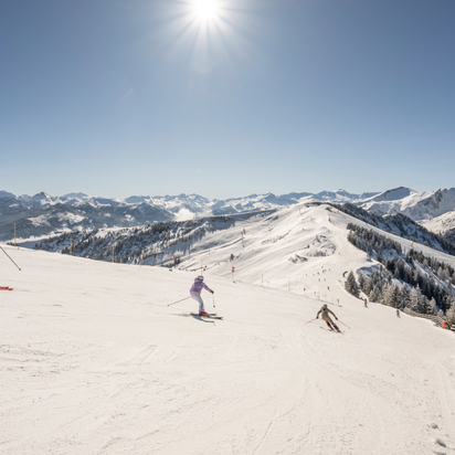 Drei Skifahrer fahren auf breiter präparierter Piste im Skigebiet Großarltal mit verschneiten Bergen unter Sonne | © Lorenz Masser