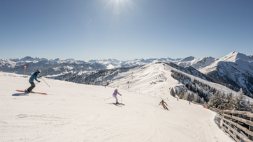 Drei Skifahrer fahren auf breiter präparierter Piste im Skigebiet Großarltal mit verschneiten Bergen unter Sonne | © Lorenz Masser