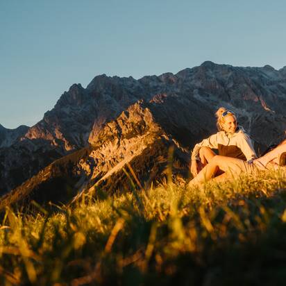 Zwei Frauen sitzen auf einer Wiese vor einem Felsmassiv in warmem Abendlicht in der Region Hochkönig. | © Hochkönig Tourismus GmbH